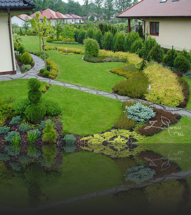 Rozległy, zadbany ogród z krętymi ścieżkami z kostki, formowanymi krzewami i rabatami z różnorodnych roślin, widok z góry na starannie zaprojektowany teren.